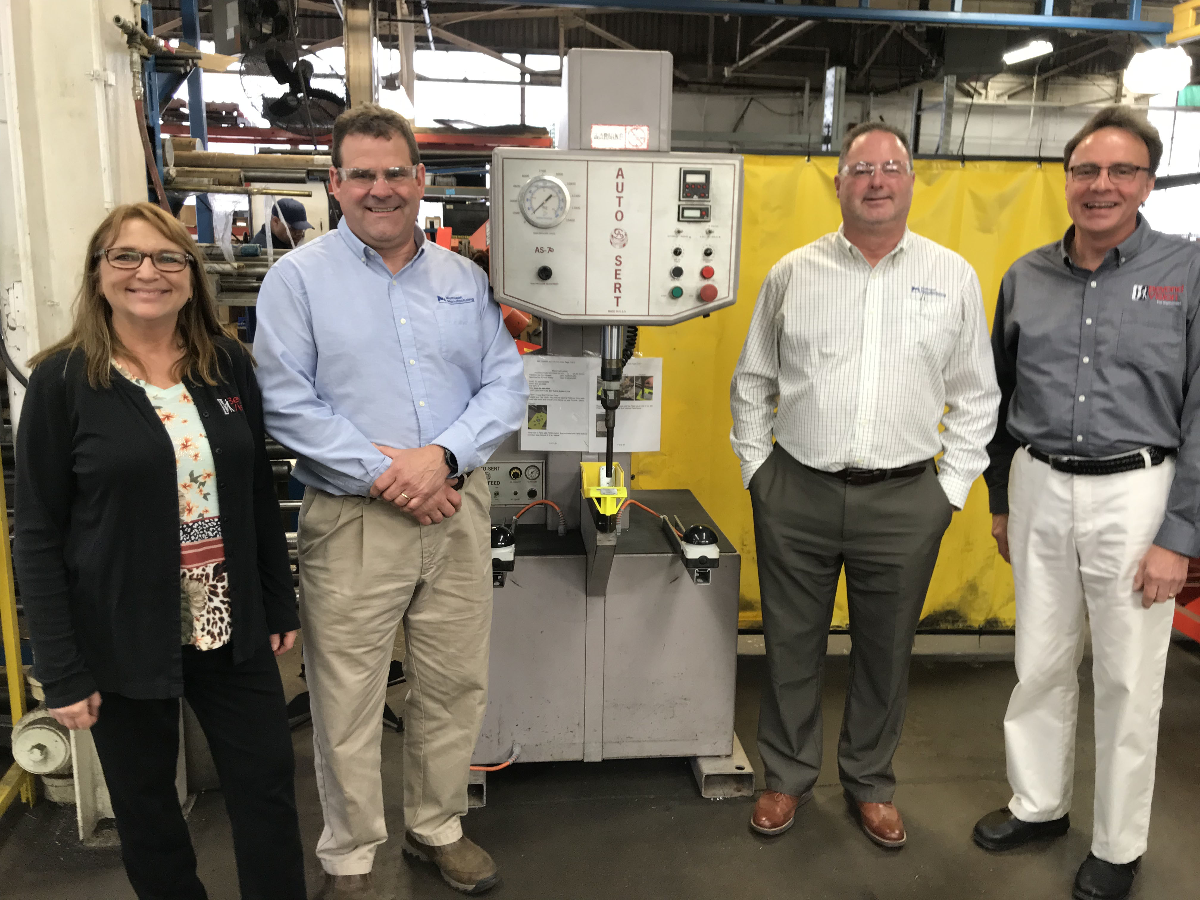 a woman and three men stand next to a narrow machine and smile. They are in a manufacturing space.