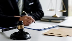 Person holding a pen and signing a document at a desk with a gavel on one side and scales on their other.